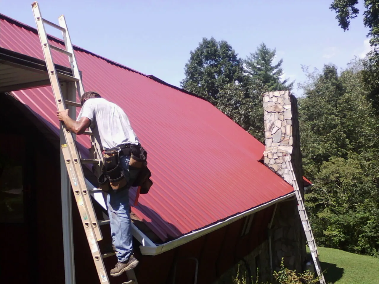 Roofer on a ladder beside a red metal roof and stone chimney — Soffit & Fascia Repair by Roofing Brothers in Muskegon